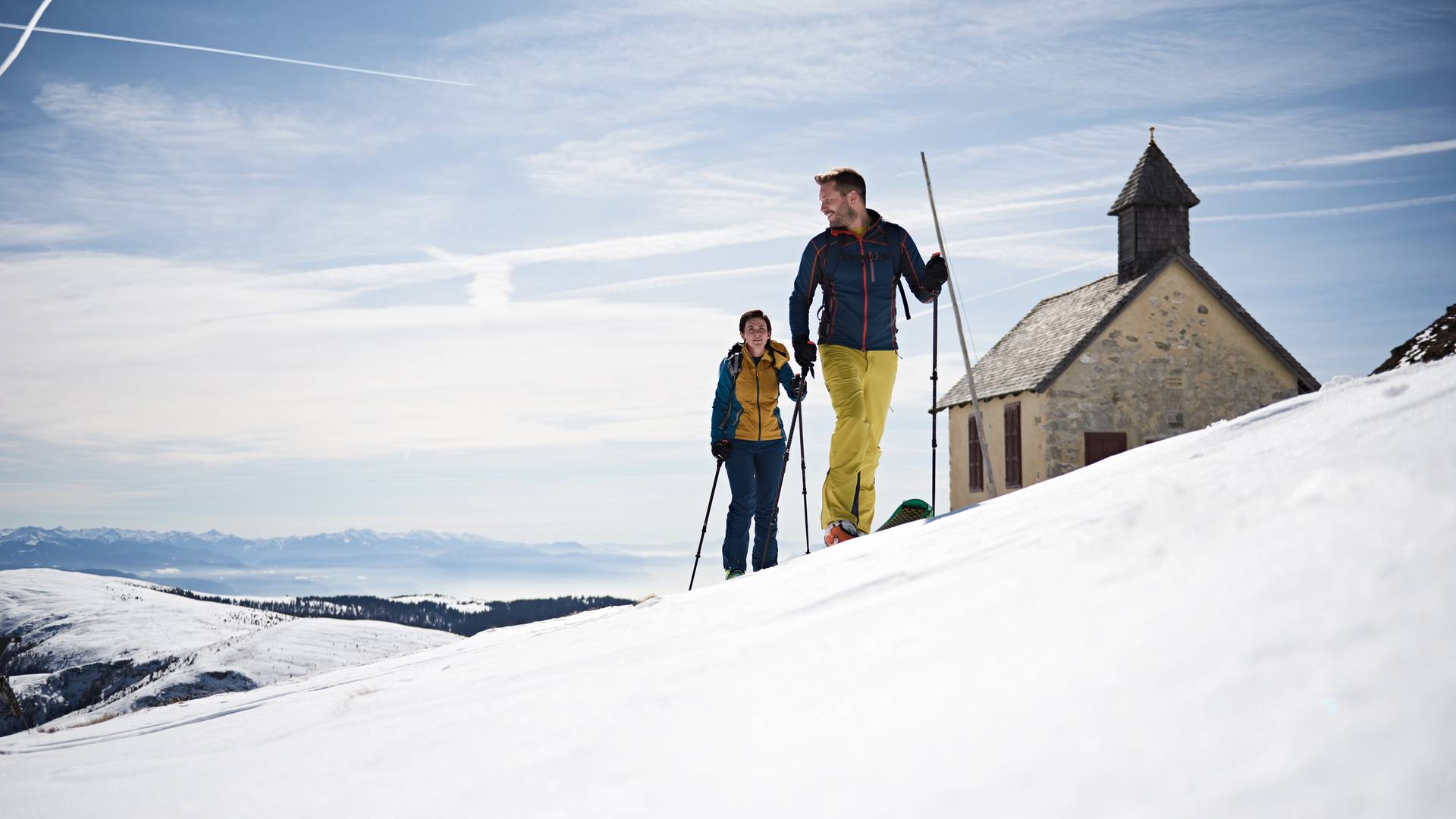 Zwei Personen beim Schneeschuhwandern vor einer kleinen Kapelle in verschneiter Berglandschaft